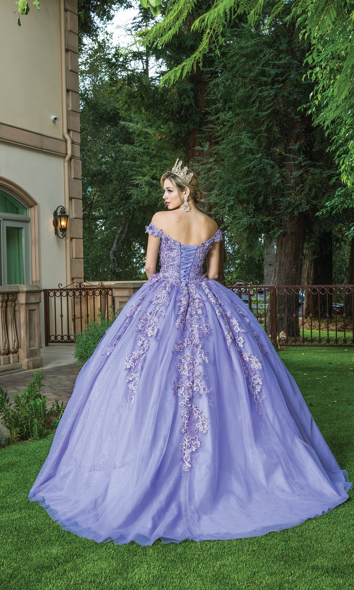 A woman in the Quinceanera Dress 1574 by Dancing Queen, featuring a sweetheart neckline and floral embroidery, stands on grass facing away near a building and tall trees.